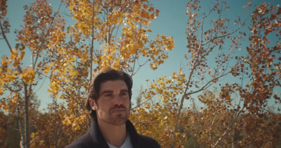 A man stands outdoors near a tree with yellowing leaves as he looks out at his surroundings during the day