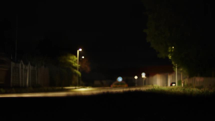 The headlights of a car illuminate grass on the side of a road as it passes by at night