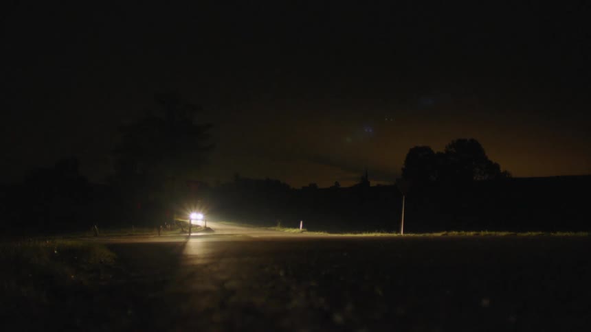 A vehicle with headlights driving on a road with trees the sky and clouds in the background
