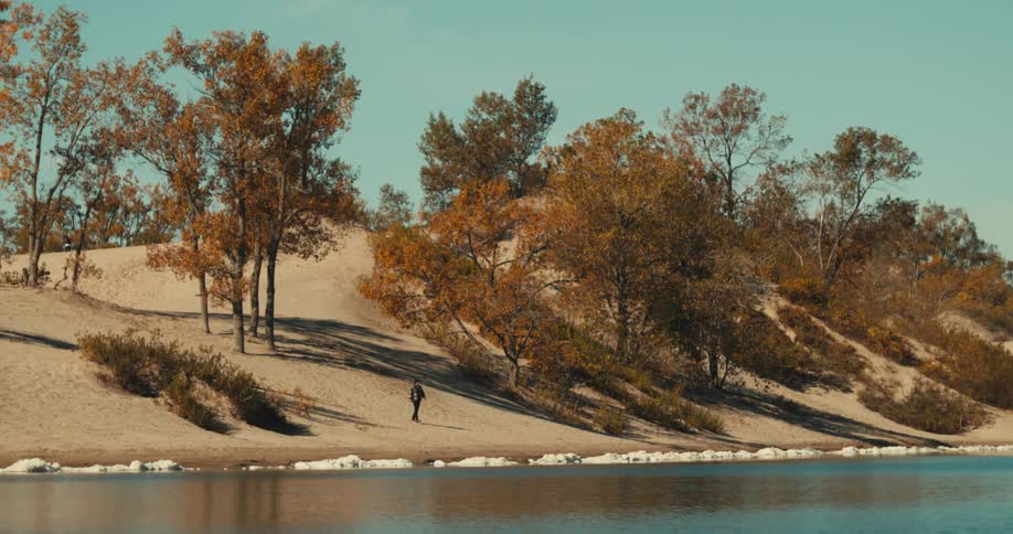 A man walks down a sandy hill towards a river lined with stones under a clear sky
