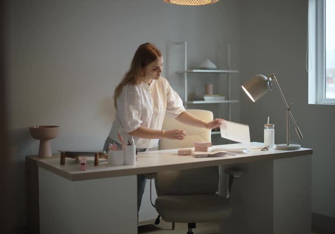 A woman places a card in a small box before placing the box inside an envelope as she stands behind a desk in an office during the day