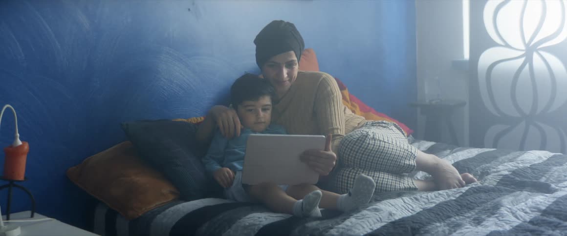 A woman curls up on a bed with her toddler son as they watch a tablet
