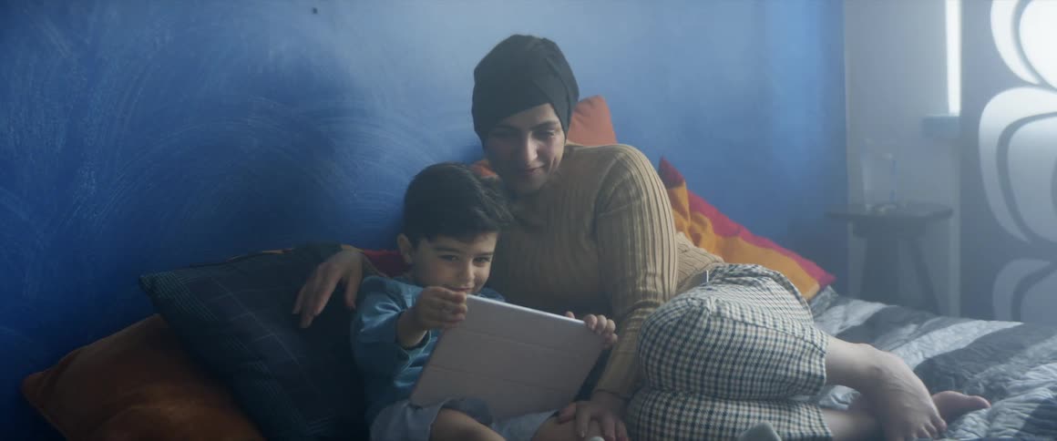 Mother and son sit on bed reading on a tablet leaning on pillows in a room with blue walls beside a table and a window