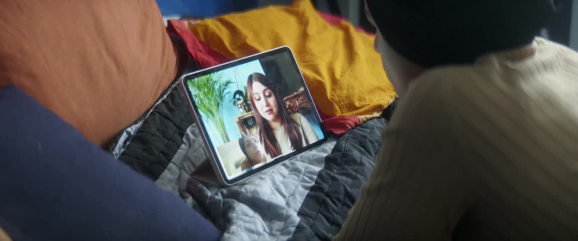 A woman lies on her bed while video chatting with another woman through her tablet