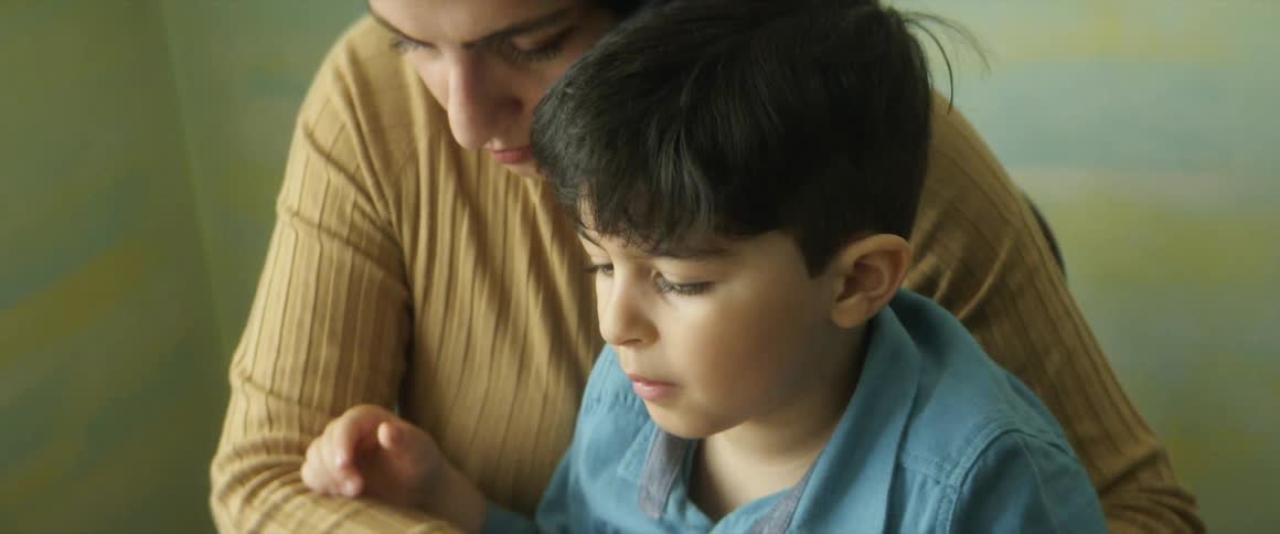 Son sits at a table and talks to a tablet computer on a table while his mother holds him