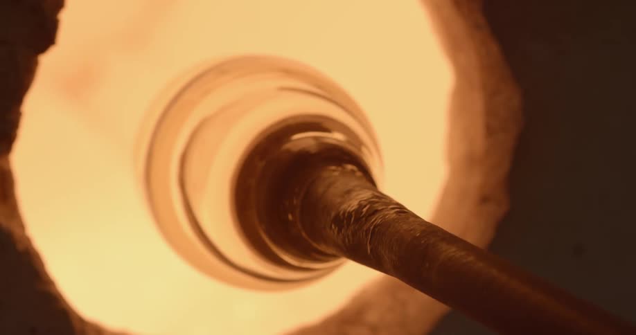 Someone turns a globule of molten glass thats hanging off of the end of a blowing rod in a kiln