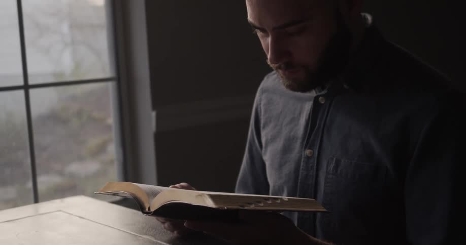 A man in a room sitting at a table reading a book with a window in the background