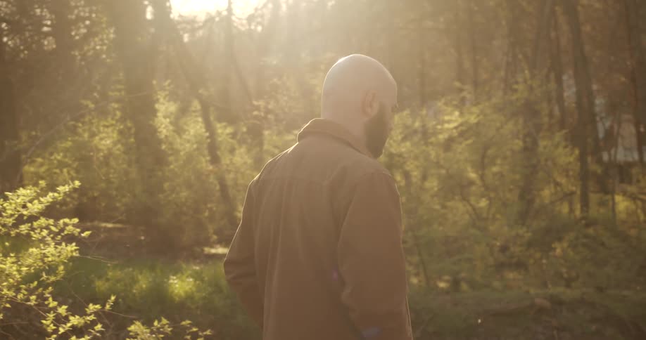 Bald man with beard walks through woods on sunny day toward buildings showing through trees