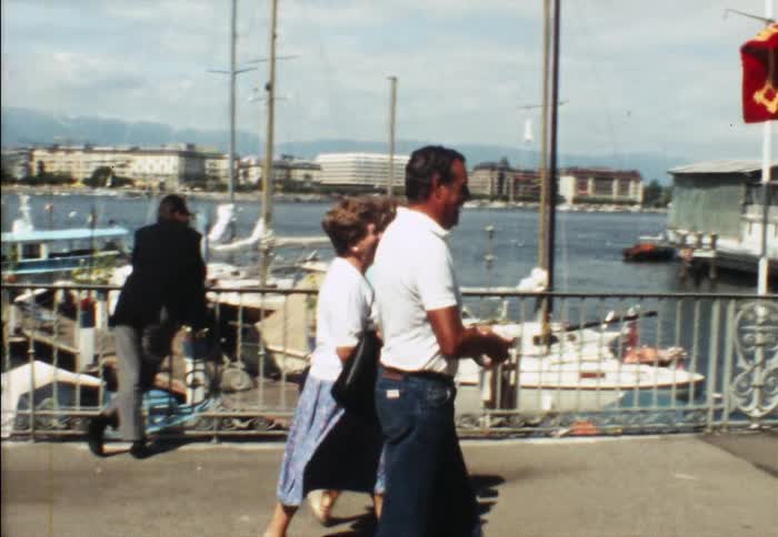 Three people walk along a pier at a marina as others lean over a railing to look at boats on the lake