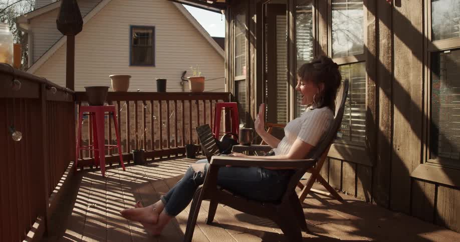 A woman sits on her backyard patio with her laptop and waves during a video call