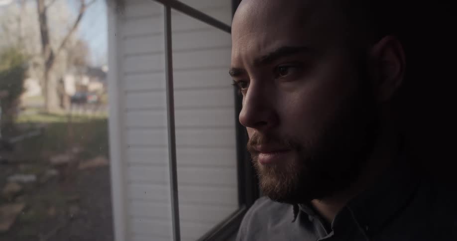 A man stares out of a window from a dark room during the day