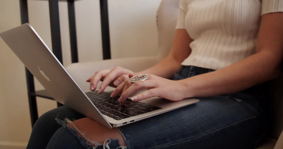 A woman sitting in a chair typing on a laptop wearing ripped jeans and a large ring