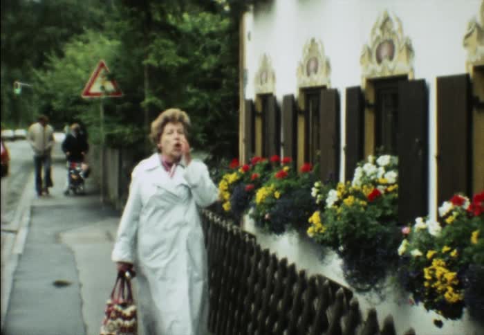 A woman blows a kiss as she walks down a sidewalk past a building with flowerbeds in windows