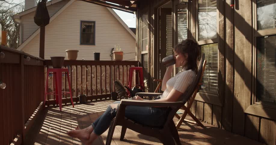 Woman sitting on balcony types on laptop in her lap and drinks from coffee mug