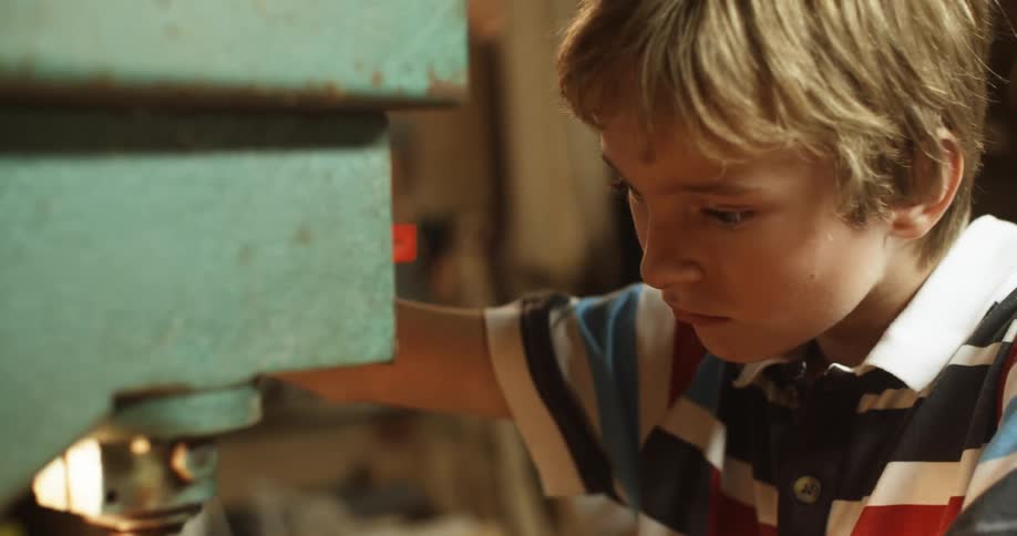 A boy uses a drill press to drill a hole in a wooden plant in a workshop