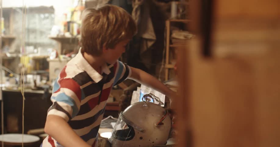 Boy working in crowded workshop attaches hose to back of wired helmet