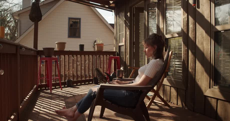 A woman sits on a chair on a porch typing on a laptop