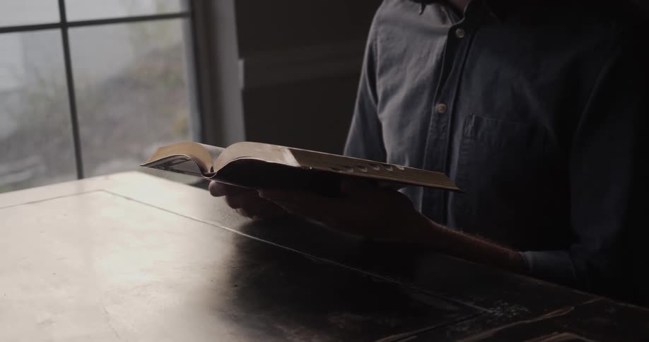 A man sitting at a table inside a dimly lit room reads a book near a window