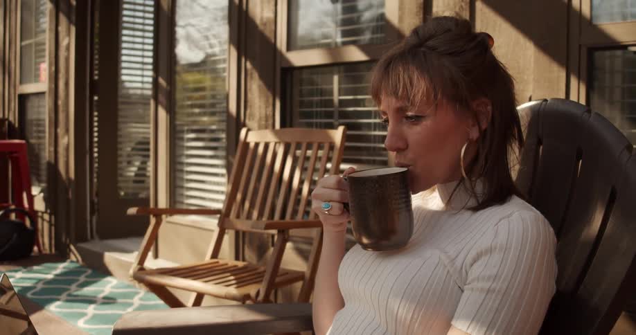 A woman on a porch sitting on a chair drinking from a mug while looking at a laptop