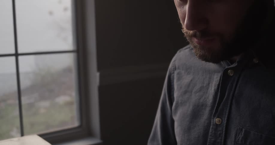 A man sitting at a table inside a dimly lit room reads a bible near a window