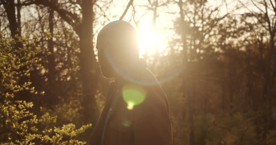 A bald man in a brown coat stands in a forest and looks around