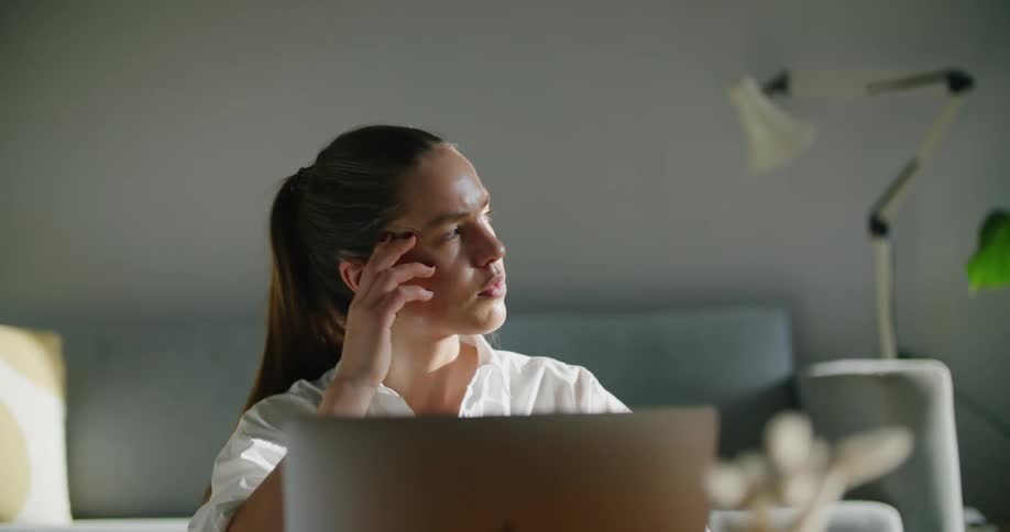 A woman sitting in front of a laptop looks off to the distance and then continues to type