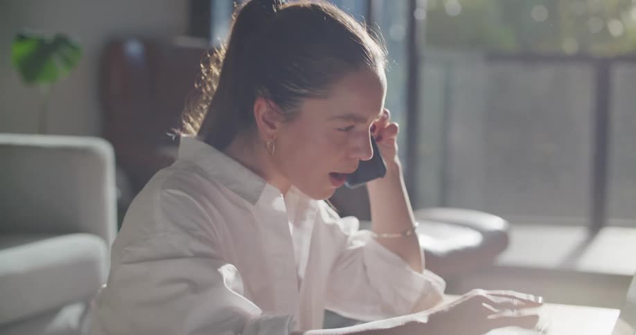 A woman sitting at her is talking on her phone while checking information on a document and on her laptop