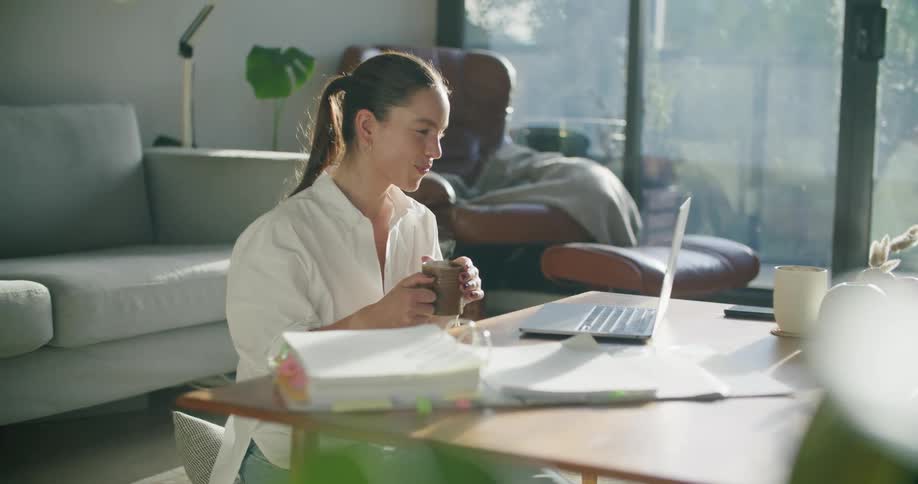 A woman sits on a pillow on the floor talking and laughing during a video call on a laptop on the coffee table