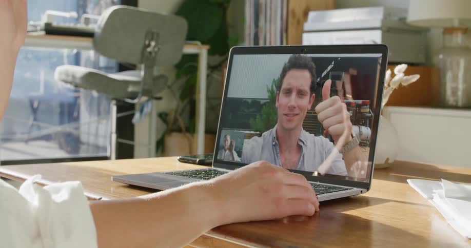 A man on a laptop screen during a video call waves and gives a thumbs up to the woman at a desk
