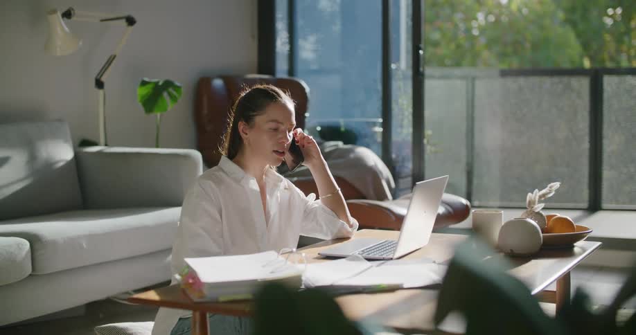 A woman sitting on the floor of her home talks on her phone while her laptop is on the coffee table in front of her