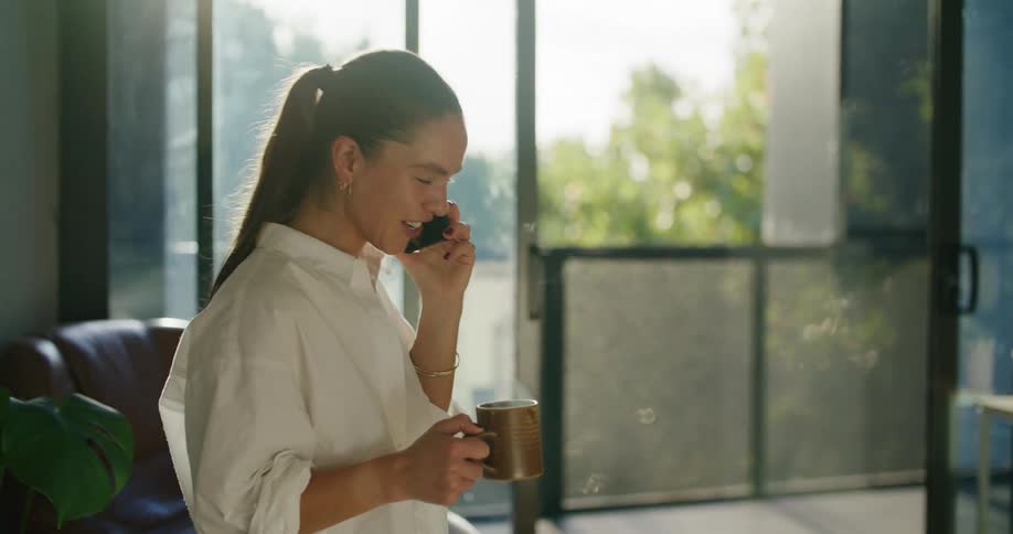 A woman laughs while holding a mug of a hot drink and talking on her phone at home