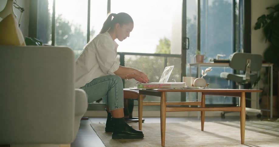 A young woman types on a laptop while glancing at the pages of a binder as she sits on a sofa in the living room of a house during the day