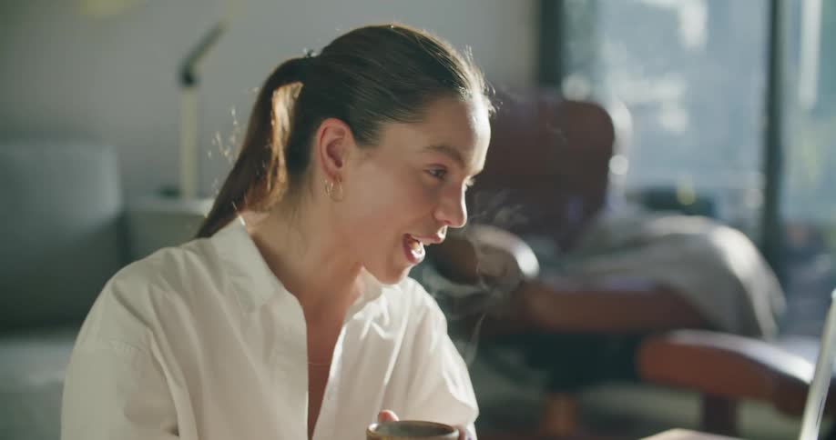 Woman sitting in the living room drinking a hot beverage while talking on a video call in a laptop