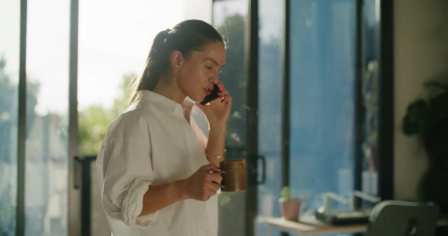 Young woman talking on her phone while drinking a hot beverage in the living room