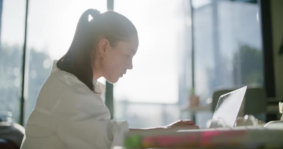 A woman sits at a desk a looks over at a document while typing on her laptop computer