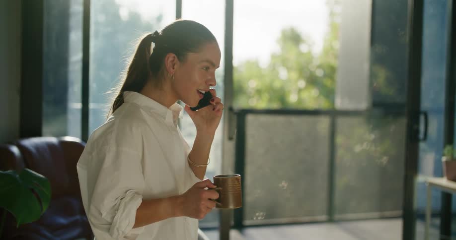 A woman in a room talking on a smart phone while holding a mug