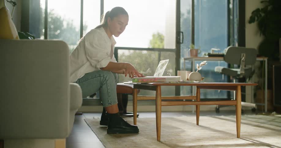 A woman sitting in a couch in the living room reads documents while using a laptop