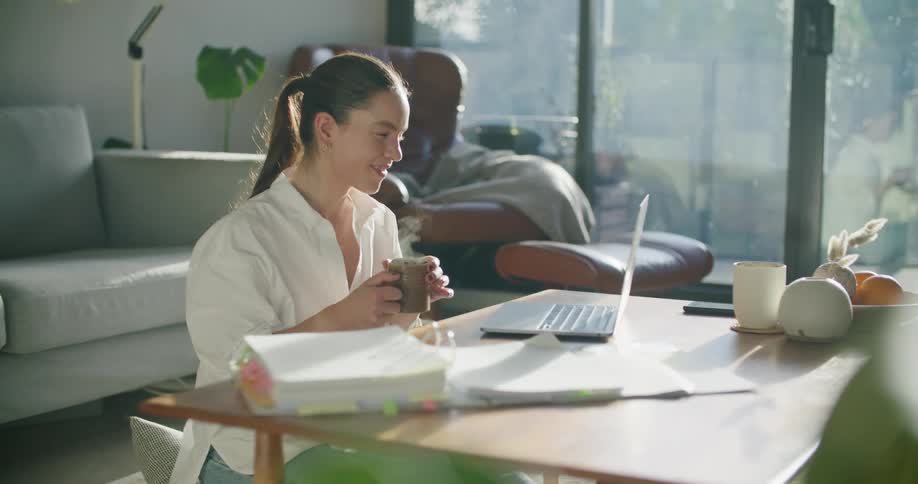 Woman sitting in the living room drinking a hot beverage while talking on a video call in a laptop