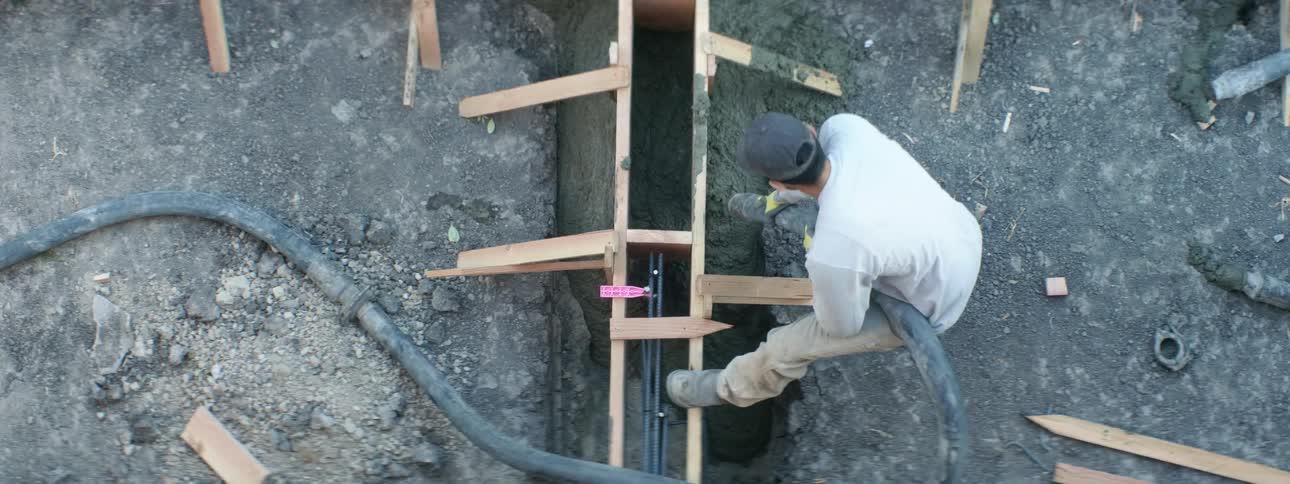 Man guides hose pouring concrete into trench around wooden frame