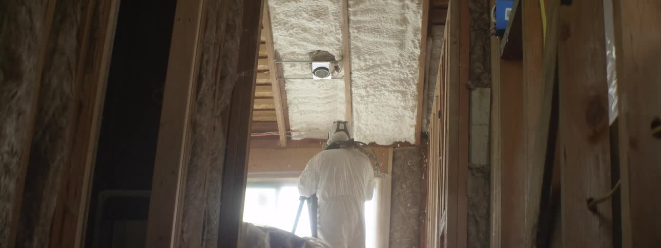 A person in a hazmat suit is spraying insulation on the ceiling beams
