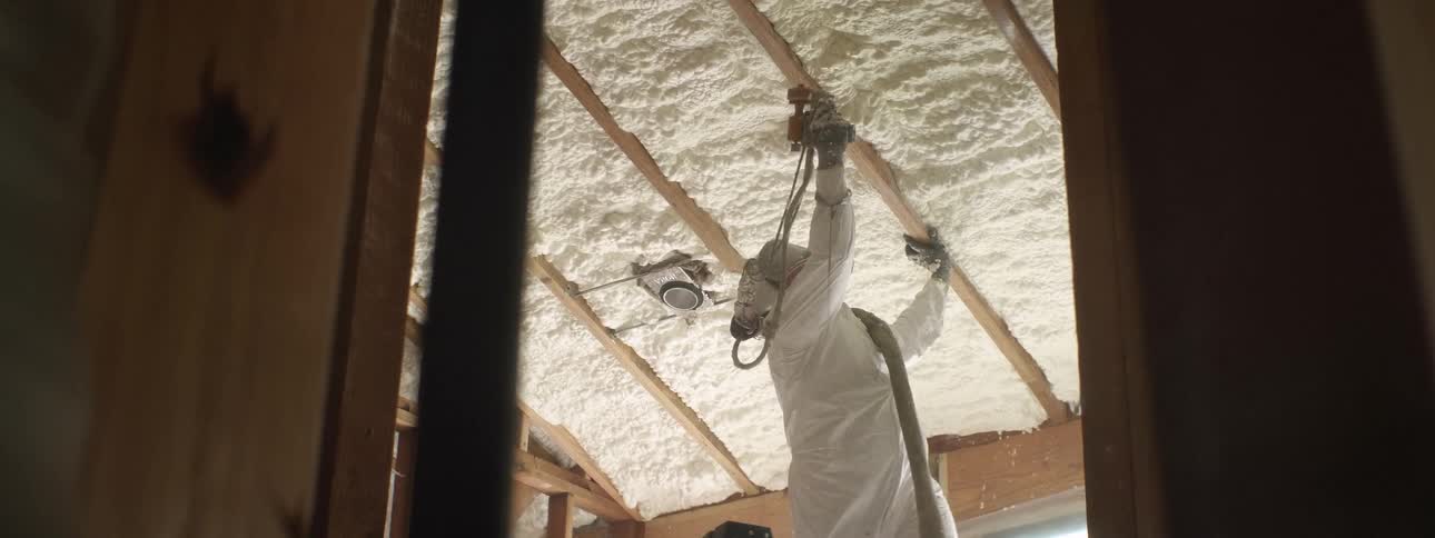 A man wearing a respirator mask and full body ppe suit steps down on a ladder after spraying foam insulation onto a newly framed ceiling