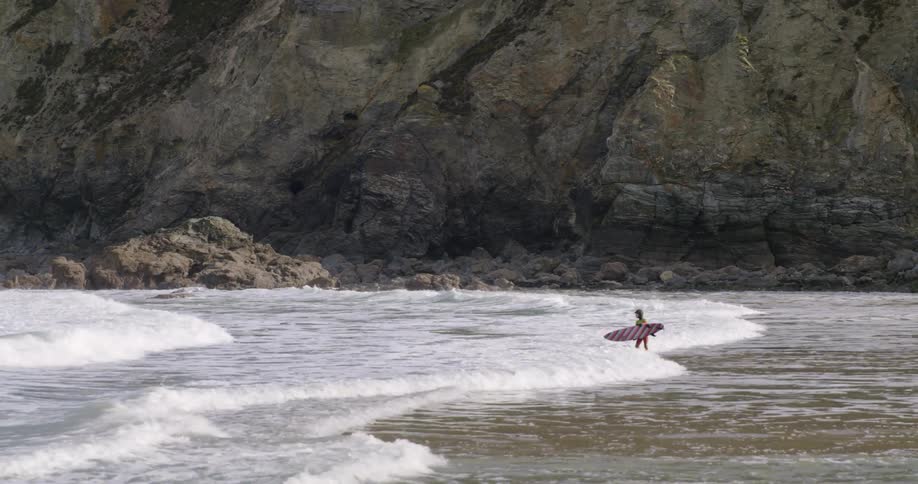 A youth with a surfboard standing on a beach with waves washing up on shore