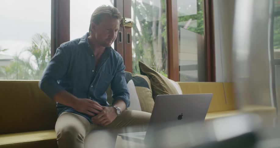 A man sitting inside on a bench waves at someone he is video chatting on a laptop