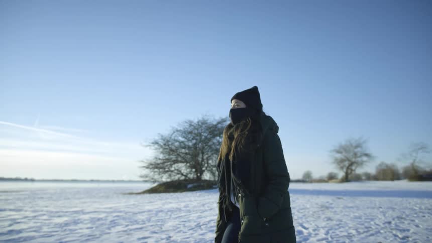 A woman walks on flat land covered in snow while wearing a mask on a sunny day