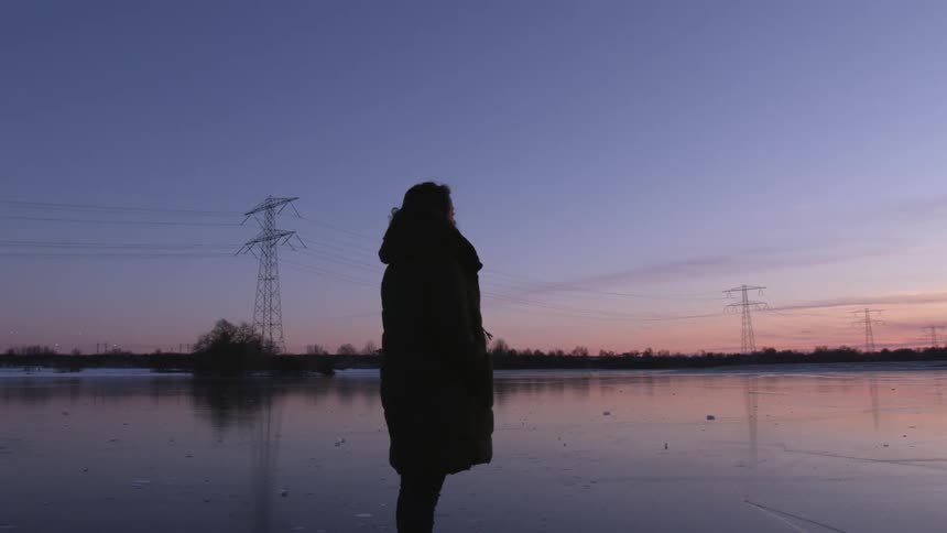 A woman wearing a coat standing near power lines and transmission towers looking at sunset