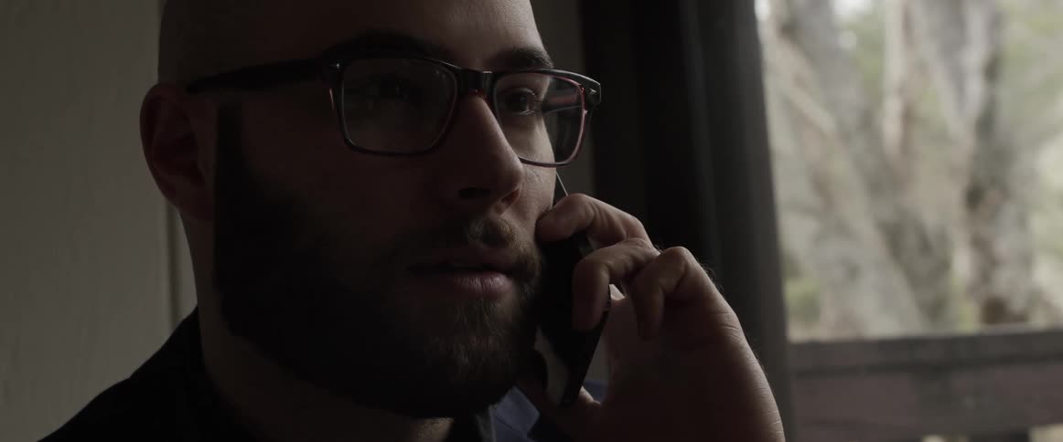 A bearded man talking on the phone inside a dimly lit room