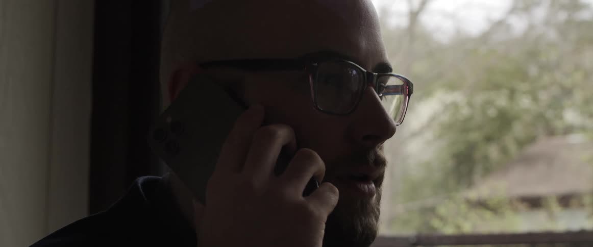 Man talks on mobile phone next to window showing neighboring house and trees