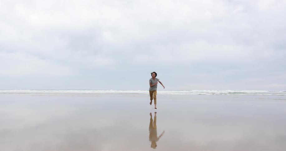 Boy with long hair runs on wet sand beach on a cloudy day