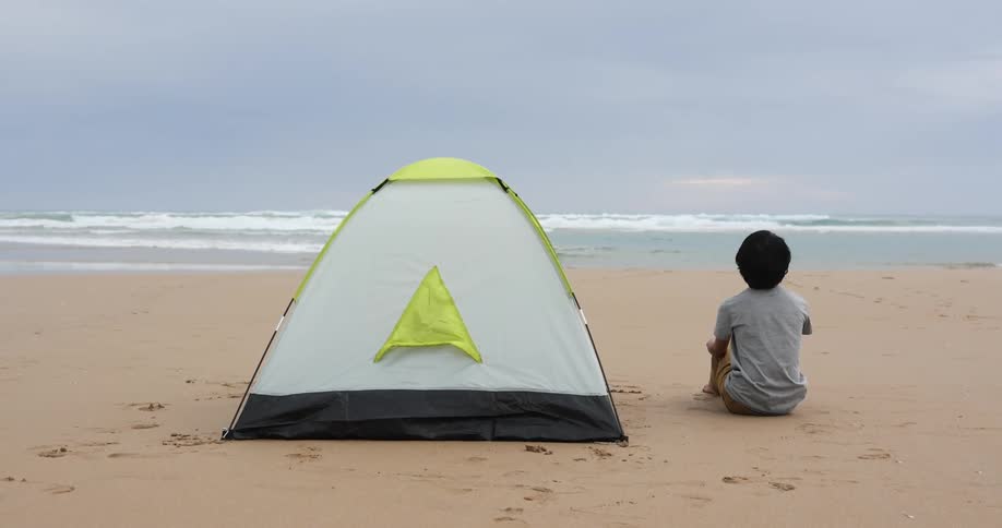 Teenage boy sits on the beach next to a tent and watches the ocean