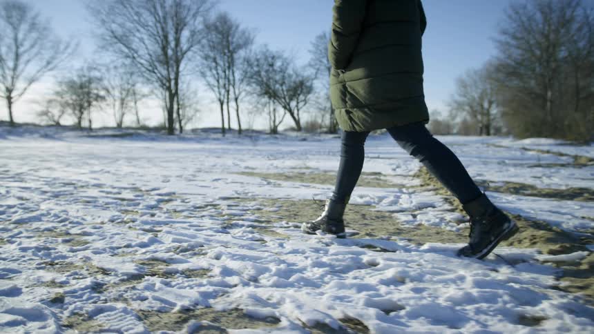 A woman in a coat and snow boots walking over a snowy field on a cold day